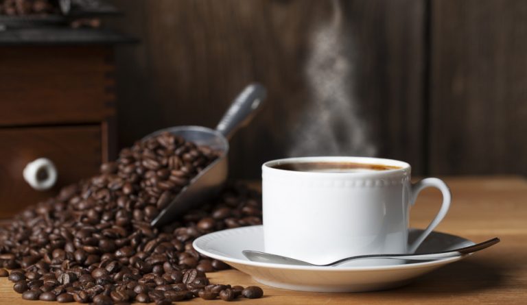 A steaming hot cup of coffee sits on a wooden table next to a metal scooper filled with coffee beans and an old fashioned coffee grinder.