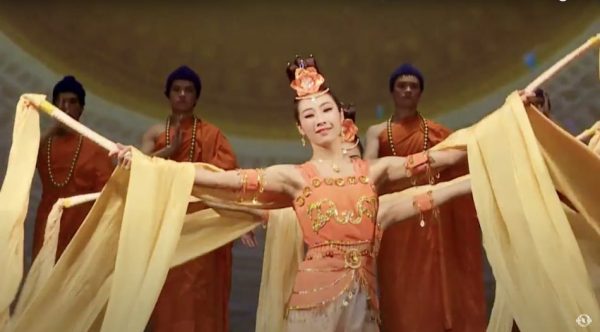 shen yun dancer in orange and yellow costume with long yellow sleeves and headpiece and male dancers dressed in orange robes with blue hair in the background.